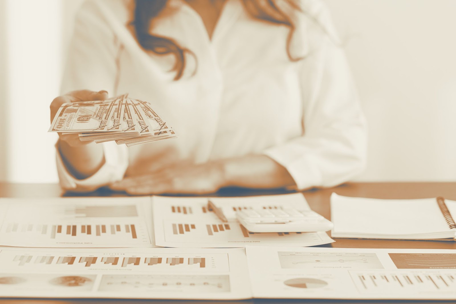 Woman holding fanned-out cash over a desk covered with financial reports and a calculator